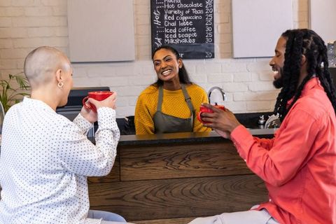 Indian barista smiling serving two customers with red mugs at cozy wood coffee counter
