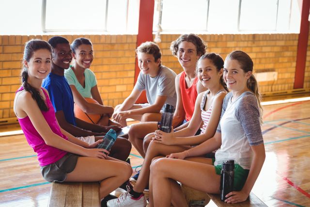 Group of high school students sitting on bench in basketball court, smiling and holding water bottles. Ideal for use in educational materials, sports promotions, youth programs, and advertisements focusing on teamwork and fitness.