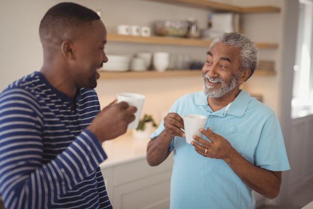 Father and son smiling and enjoying coffee together in a modern kitchen. Perfect for use in family-oriented advertisements, lifestyle blogs, and articles about family bonding, morning routines, or multigenerational relationships.