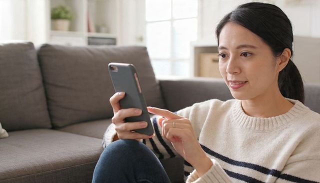 Asian woman scrolling smartphone on sofa in sunlit living room, cozy home lifestyle