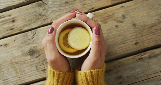 Image of hands of caucasian woman holding mug with tea and lemon on wooden surface. seasons, autumn, coziness and relax concept.