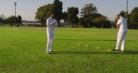 Two cricketers practicing bowling drill on sunlit club outfield with yellow cones