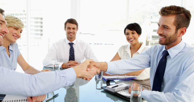 Group of business professionals sitting around table, two men in foreground shaking hands symbolizing agreement or partnership, others smiling. Useful for concepts like business collaboration, success, teamwork, formal meetings, corporate agreements.