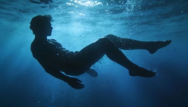 Floating male silhouette beneath sunbeams in deep blue water, serene aquatic solitude