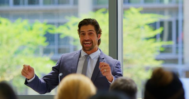 A charismatic public speaker in a suit is energetically engaging with an audience in a modern office setting. His expressive gestures and enthusiastic demeanor captivate the audience's attention. This image can be used in articles about public speaking tips, leadership seminars, business communication skills, or promotional materials for corporate events.