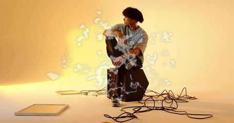 Contemplative musician sitting on amp listening with earphones among cables and warm glow