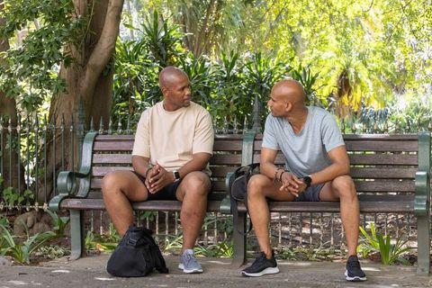 Two african american men chatting on park bench in casual activewear, urban green space