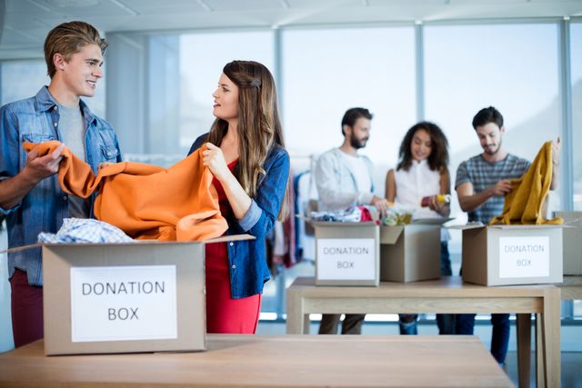 Group of colleagues sorting clothes into donation boxes in a modern office. Ideal for use in articles or campaigns about corporate social responsibility, community service, teamwork, and charitable activities.