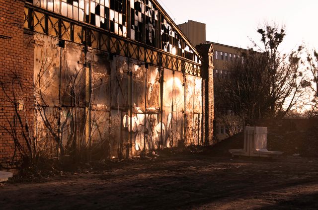 Exterior of abandoned warehouse featuring rusty, dilapidated walls in urban area during sunset. Graffiti present on structure, overgrown vegetation surrounding. Can be used for themes of urban decay, abandoned places, post-apocalyptic, or photography with an industrial backdrop.