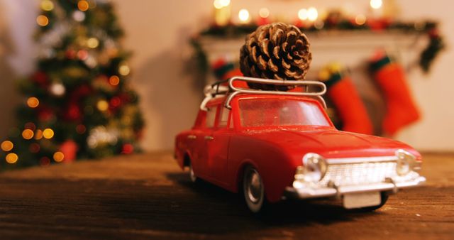 This image depicts a vintage red toy car carrying a pinecone on its roof, with warm Christmas decorations in the background. A Christmas tree adorned with ornaments stands on the left, while candles and stockings are hung on a mantelpiece in the background. Ideal for holiday greeting cards, festive advertising campaigns, and Christmas decoration promotions.