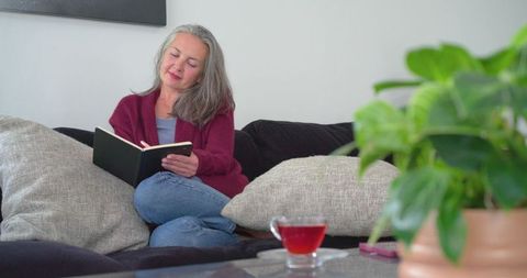 Mature woman relaxing on sofa reading book while sipping tea in cozy home