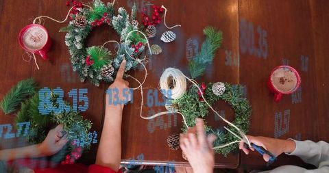 Women crafting holiday wreaths at wooden table with twine, pinecones, berries and hot cocoa