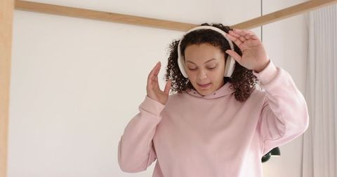 African american woman adjusting headphones in cozy modern bedroom wearing pink hoodie