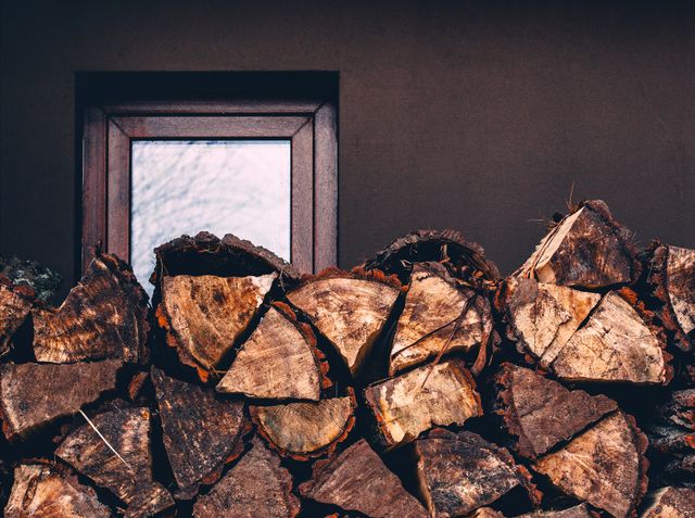 Wood logs neatly stacked in front of a wood cabin window, creating a warm and rustic atmosphere. Ideal for illustrating themes related to outdoor living, rural life, home heating, or firewood preparation. Could be used in magazines, blogs, or advertisements focusing on country living, sustainable practices, or winter preparation.
