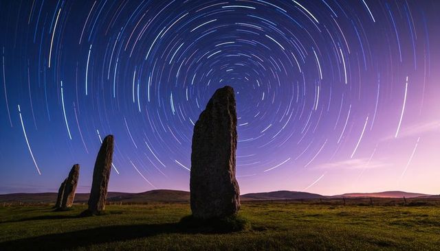 Ancient standing stones under circular star trails over purple twilight moorland