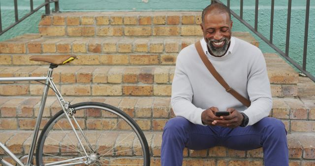 Man is sitting on steps in a casual setting with a bicycle nearby, engaged with his smartphone and smiling. The setting appears relaxed, suggesting leisure and easy-going moments. Suitable for illustrating themes of technology use, leisure, lifestyle, and happiness.