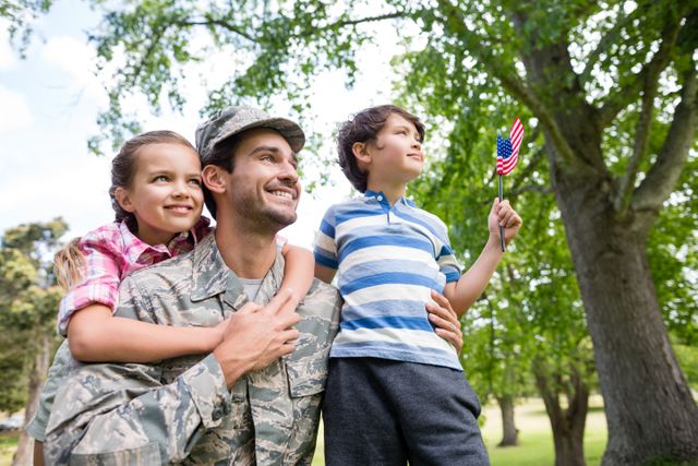 Soldier in uniform enjoying a sunny day in the park with his son and daughter. The boy holds an American flag, symbolizing patriotism. Ideal for use in content related to military families, reunions, patriotism, and family bonding.