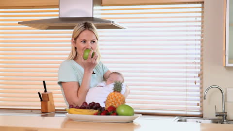 Mother Enjoying Healthy Snack While Holding Sleeping Baby in Kitchen
