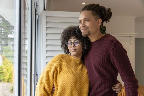 African american couple standing by glass door wearing knit sweaters looking out at garden