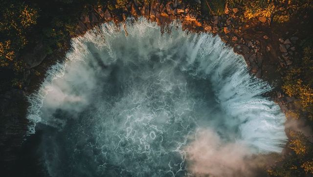 Aerial surf crashing against rocky cove with teal water and rising mist