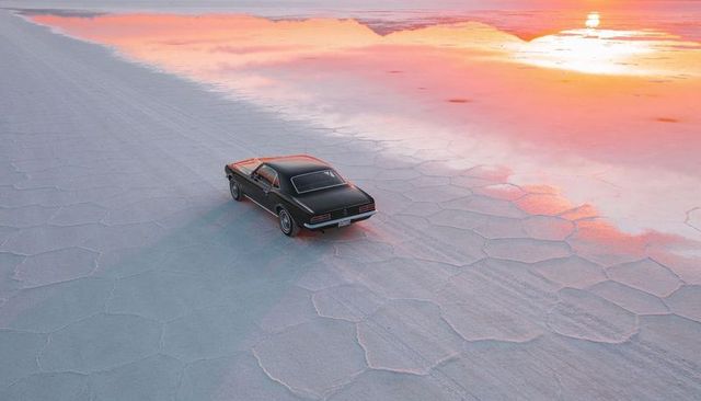Driving black muscle car across salt flats sunset with pink sky reflection and tire tracks