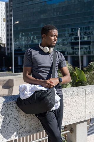 African american man leaning on overpass railing wearing headphones and crossbody bag