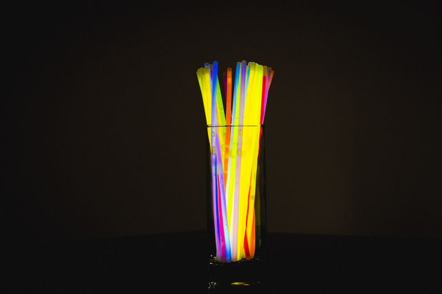 Illuminated multicolored neon drinking straws in a glass against a black background. The vibrant colors and glowing effect make this image ideal for use in party invitations, nightlife promotions, festive decorations, and abstract art projects. The dark background enhances the brightness of the neon straws, creating a striking visual contrast.