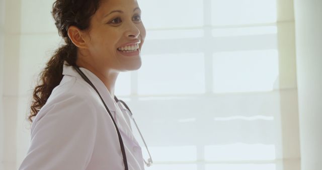 A smiling young female doctor with a stethoscope around her neck stands confidently, with copy space. Her cheerful demeanor and professional attire suggest a positive healthcare environment.