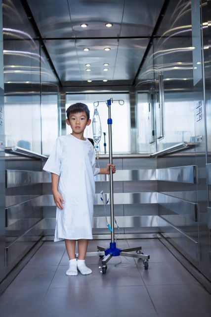 Young boy patient standing in a hospital elevator holding an IV drip stand. He is wearing a hospital gown and appears to be in a healthcare facility. This image can be used for healthcare, pediatric care, medical treatment, hospital environment, and recovery-related content.