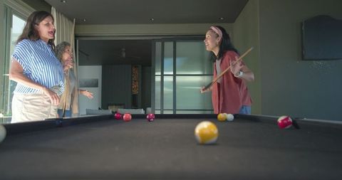 Three women playing pool around modern table laughing and talking in contemporary home