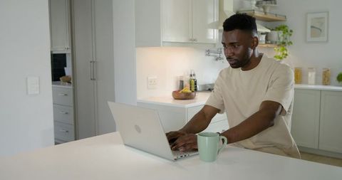African man working on laptop at modern white kitchen island with green mug