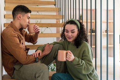 Diverse couple relaxing on wooden stairs holding matching mugs in cozy modern interior