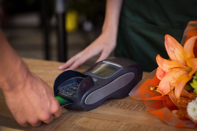 Customer swiping credit card at point of sale terminal in flower shop. Ideal for illustrating retail transactions, customer service, and small business operations. Useful for articles on payment methods, retail technology, and shopping experiences.