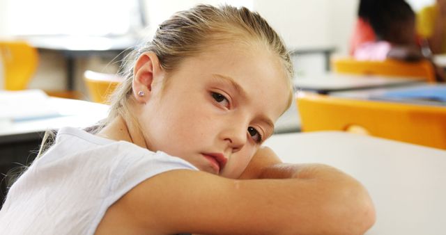 Young girl with a pensive expression leaning on a desk in a classroom. Ideal for educational materials, articles on children's emotional well-being, or school-related websites featuring student engagement and classroom environments.