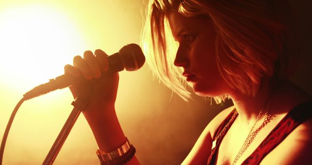 A young Caucasian woman is performing as a singer, with a microphone in hand and dramatic lighting enhancing the mood. Her intense expression and the warm stage lighting suggest a passionate musical performance.