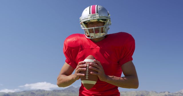 Football player in red jersey holding football, standing against clear blue sky outdoors. Useful for sports ads, fitness campaigns, football academy promotions, and articles about sports and athletics.