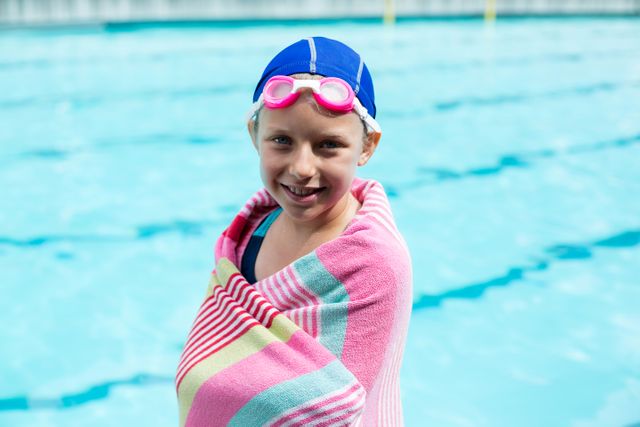 Young girl standing by the pool wrapped in a colorful towel, smiling after swimming. Ideal for use in advertisements for swimming lessons, summer camps, children's sports activities, or healthy lifestyle promotions.
