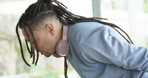 Young man with dreadlocks working on laptop wearing pink headphones in sunlit workspace