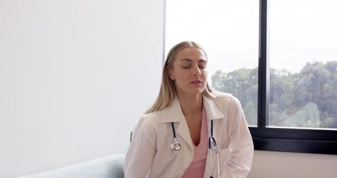Female clinician resting with eyes closed, sitting by window wearing white coat and stethoscope