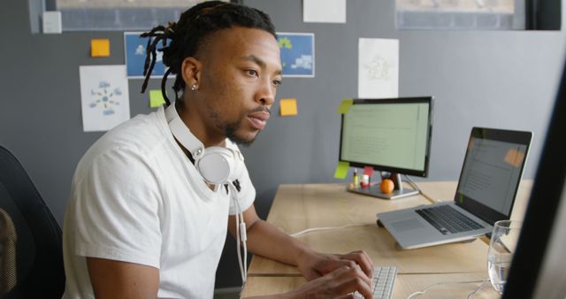 Young man focused on work at a desk in a modern office setting. He uses a computer and laptop with headphones around his neck, indicating a high-tech and busy work environment. Great for depicting corporate work, tech industry jobs, millennials in the workplace, and modern office dynamics.