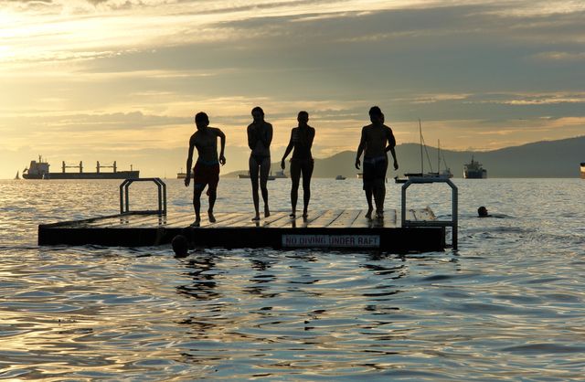 Group of friends standing on a floating dock during sunset, enjoying summer vacation by the ocean. Silhouetted figures create a sense of adventure and camaraderie. Suitable for travel advertisements, vacation brochures, lifestyle blogs and outdoor activity promotions.