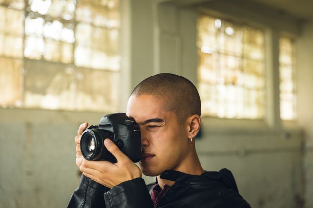 Side view close up of a hip young biracial man in an empty warehouse, taking photos with SLR camera, wearing black jacked.