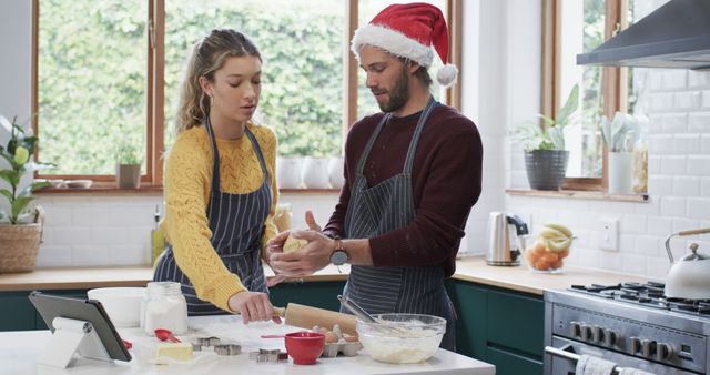 A couple is baking pastries together in a modern kitchen during the holiday season. Both are wearing aprons, and the man is wearing a Santa hat. They are working together with ingredients and a tablet is seen on the counter. This image is perfect for advertisements, holiday articles, cooking blogs, and promotions related to family bonding, festive preparation, and home cooking.