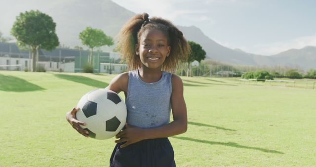 Young African American girl holding soccer ball in a park on a sunny day. She is smiling and appears happy, exemplifying an active and joyful childhood. Ideal for use in advertisements promoting outdoor activities, sports events, children's programs, health and fitness campaigns, or summer recreation.