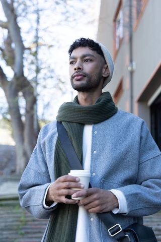 Young man holding coffee on urban walkway wearing grey coat and green scarf