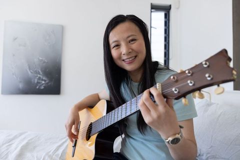 Asian woman playing acoustic guitar on bed, smiling and relaxed, home musician lifestyle
