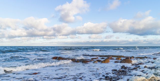 Serene scene of a rocky seashore with gentle waves lapping against the shore and fluffy clouds scattered across a blue sky. Perfect for travel websites, nature scene illustrations, relaxation and meditation themes, beach vacation promotions, and environmental campaigns.