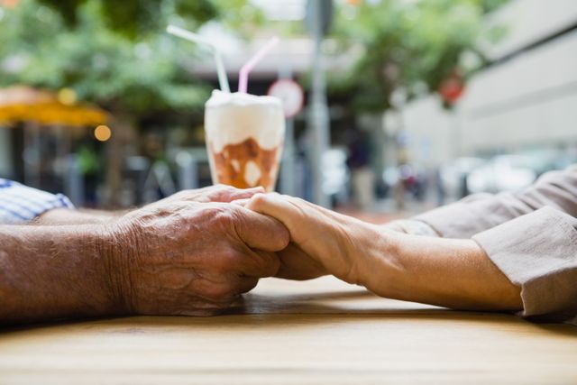 Elderly couple holding hands across a table in an outdoor cafe, with a milkshake in the background. Perfect for themes related to romance, senior love, and relationships. Great for use in advertisements, greeting cards, and social media posts focusing on love, connection, and affection among the elderly.