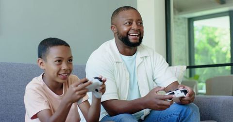 Father and son playing video games on couch, laughing and bonding in bright living room