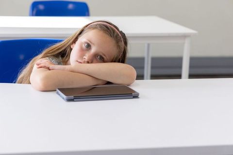 Thoughtful schoolgirl resting on desk with closed tablet in bright empty classroom awaiting lesson